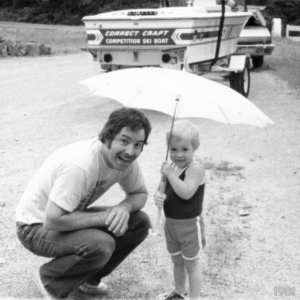 Gary Breisch with his son, Robert Breisch, in 1981 in the rain, enjoying being. That's the blue and white ski boat and 1974 El Camino in background.